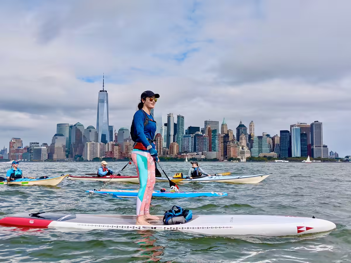 A woman riding a SIC RS stand-up paddleboard with a group of sea kayakers heading toward Surf City Restaurant. The lower Manhattan skyline, including One World Trade, are visible across the Hudson River.