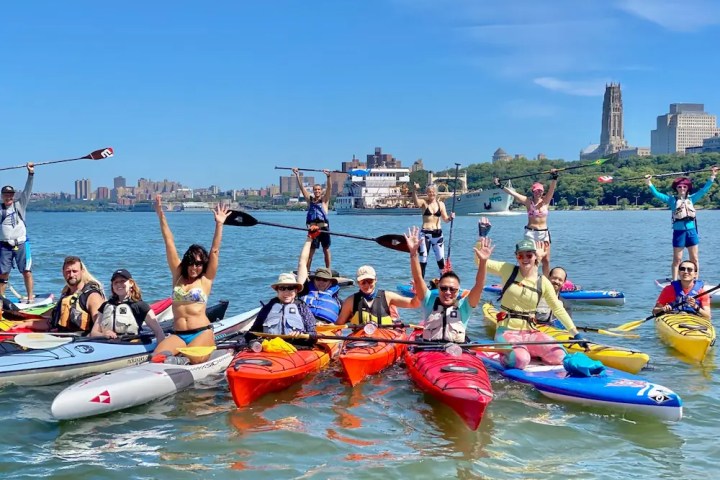 A group of 15 smiling Adventure Club members raising their paddles in the air on a sunny summer day in New York City.