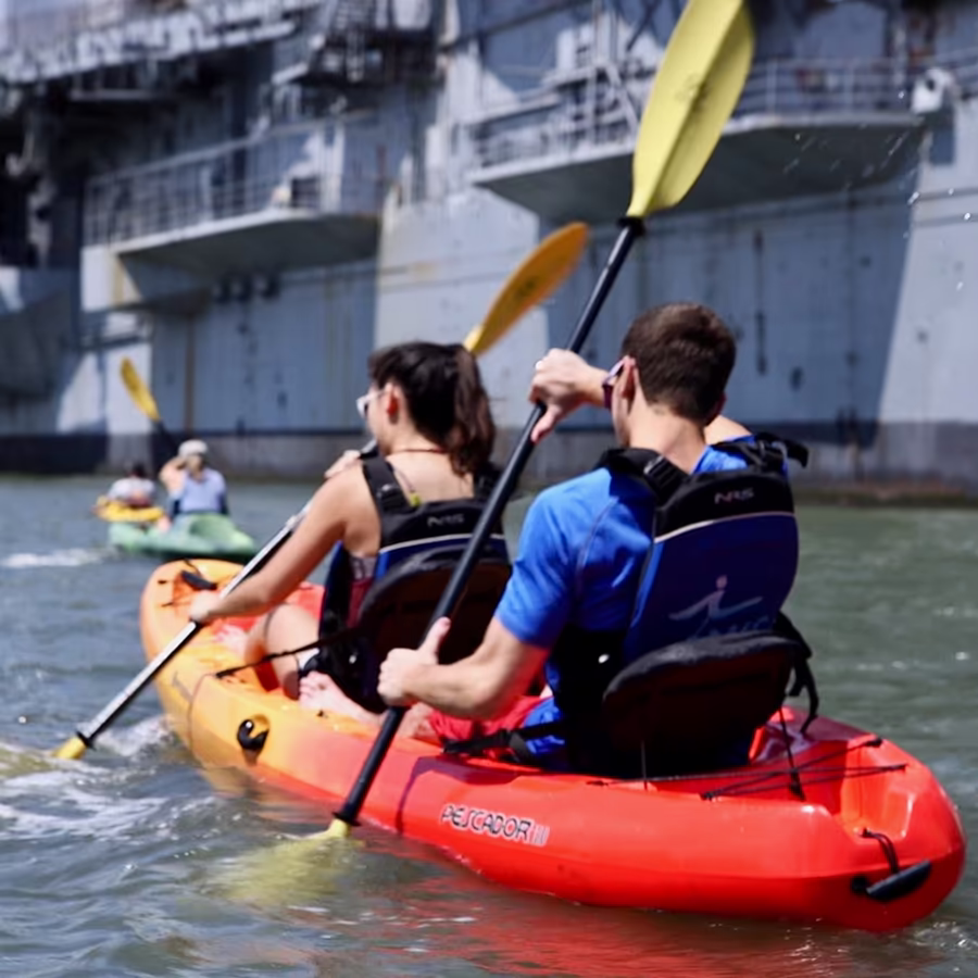 A young couple paddling a tandem kayak rental together in the Hudson River with the Intrepid Museum in the background. Active date idea and sightseeing at Manhattan Kayak, Pier 84, NYC.