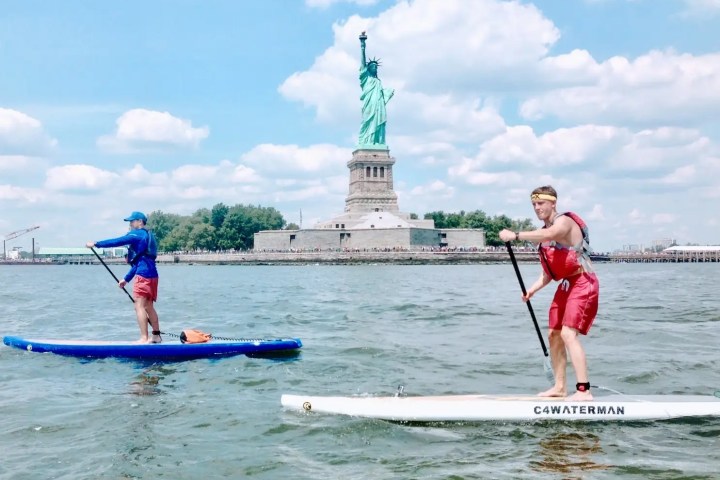 Two men stand up paddleboarding at the Statue of Liberty on a summer day.