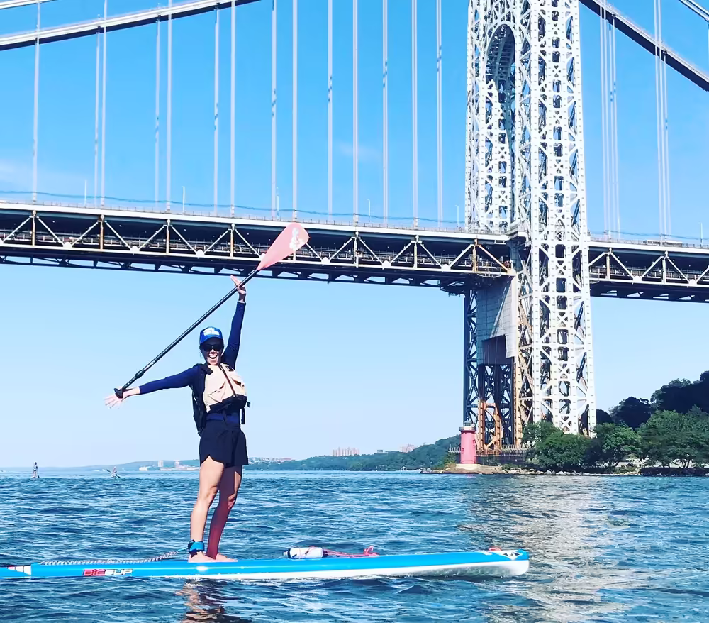 On a bright blue summer day, a woman on a stand-up paddleboard raises her paddle into the air just in front of the George Washington Bridge on the Hudson River..