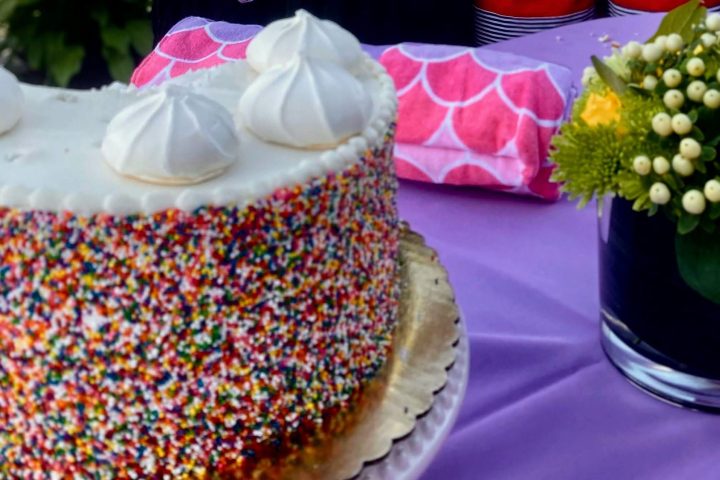 A birthday cake with rainbow sprinkles on an outdoor table with a purple tablecloth.