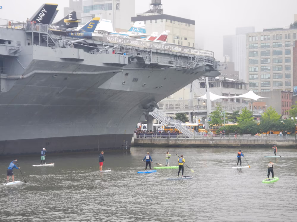 A Paddleboard Basics 2 class on a calm, foggy morning in the Pier 84 cove. Two professional Manhattan Kayak instructors lead a group of seven students on colorful boards, dwarfed by the massive hull of the USS Intrepid Museum.