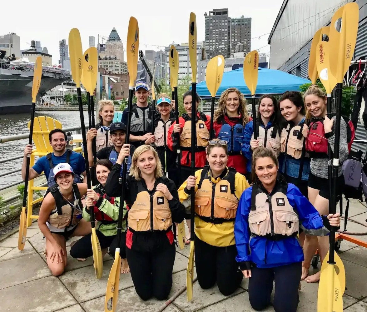 A professional NYC corporate team in gear with paddles and life jackets, ready for a private group kayak trip at a Midtown Manhattan boathouse.