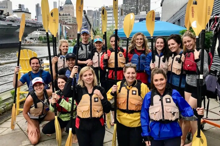 A professional NYC corporate team in gear with paddles and life jackets, ready for a private group kayak trip at a Midtown Manhattan boathouse.
