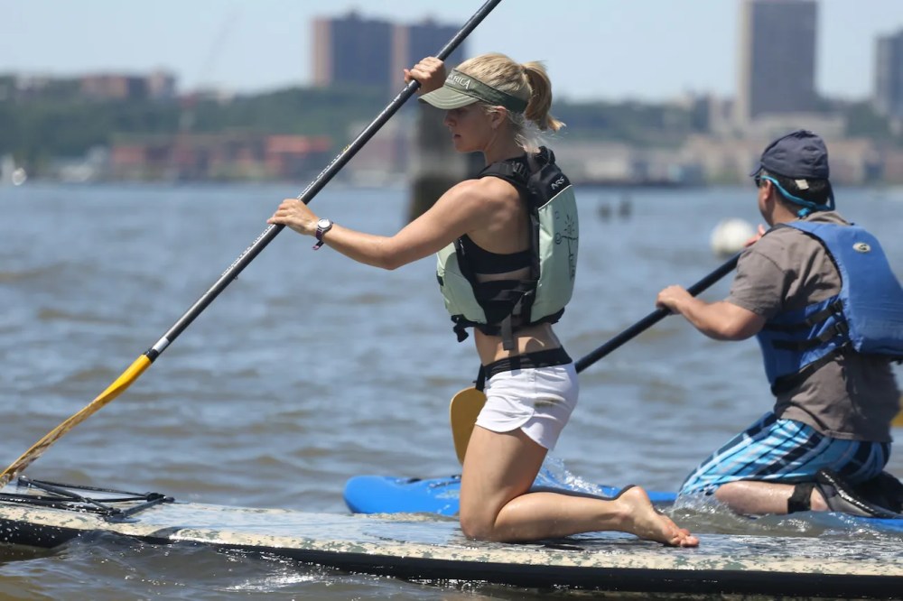 An instructor at Manhattan Kayak Co practices an upright kneeling stroke on a paddleboard, focusing on a wide paddle grip and forward gaze during a lesson.