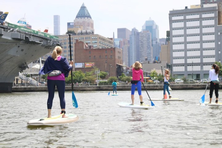 A milestone moment: A professional Manhattan Kayak instructor leads four students standing tall on their paddleboards in the Hudson River. The group faces the Midtown Manhattan skyline, with the copper-topped One Worldwide Plaza tower in the distance, during a Paddleboard Basics 2 class.
