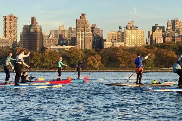 A group of Manhattan Kayak students graduate to the open river during a Paddleboard Basics 3 class. They chat and smile while paddling together on calm water, with the sunlit Upper West Side skyline and Riverside Park in the background.