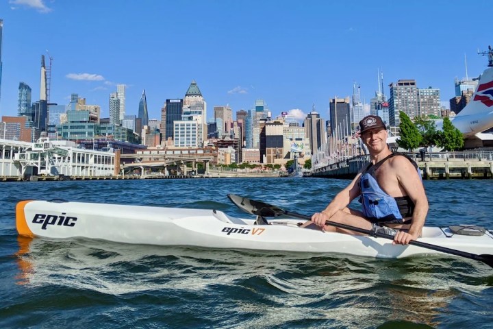 A student on a private surfski lesson navigates the Hudson River on an introductory Epic V7 surfski. He smiles during his third session on the open water, with the Midtown Manhattan skyline and the British Airways Concorde at the Intrepid Museum in the background.