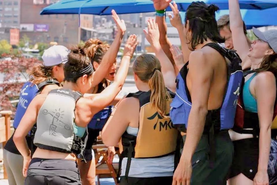 A cheering corporate team celebrating with high-fives on a waterfront patio after a successful NYC private kayak trip.
