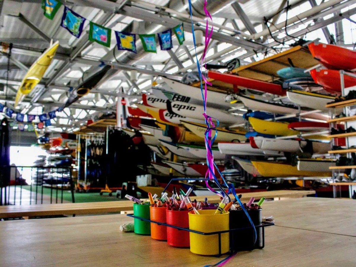 The interior of a large, professional NYC boathouse at Pier 84, featuring rows of kayaks and paddleboards, and organized activity spaces for public programming.
