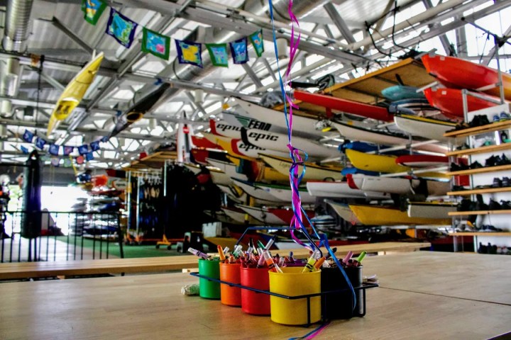 The interior of a large, professional NYC boathouse at Pier 84, featuring rows of kayaks and paddleboards, and organized activity spaces for public programming.