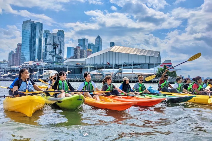 Smiling university students and interns rafting their kayaks together on the Hudson River with the USS Intrepid and Concorde in the background.