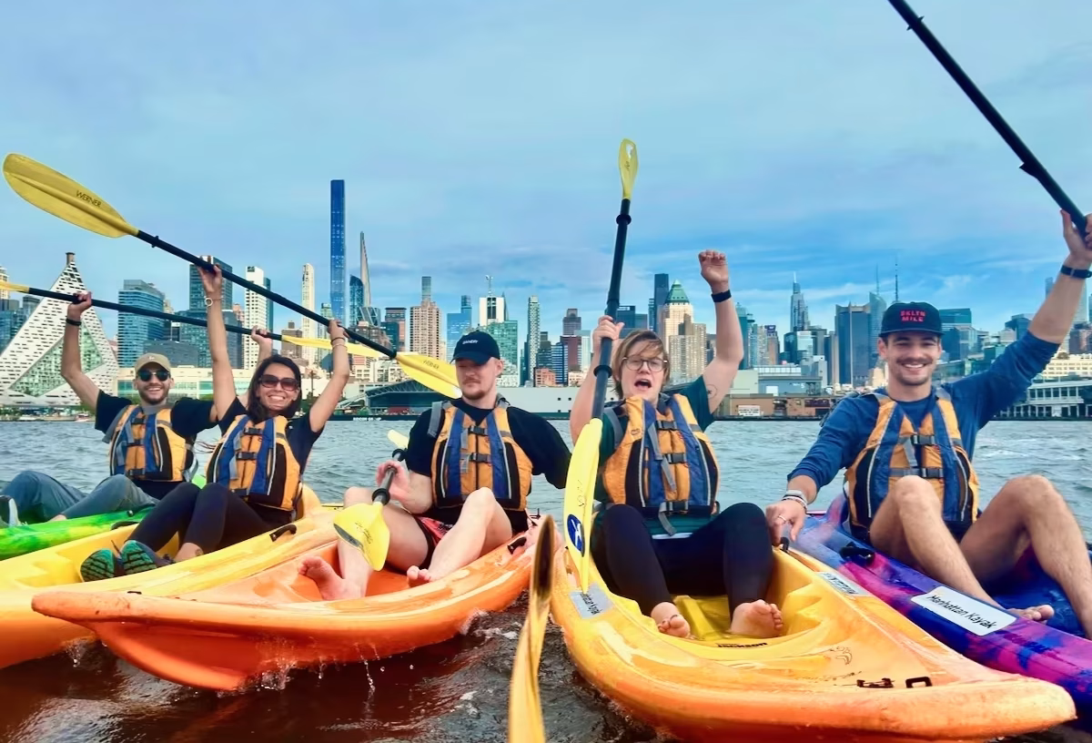 A small team summer outing on the water with raised paddles against the backdrop of the iconic NYC skyline and skyscrapers.