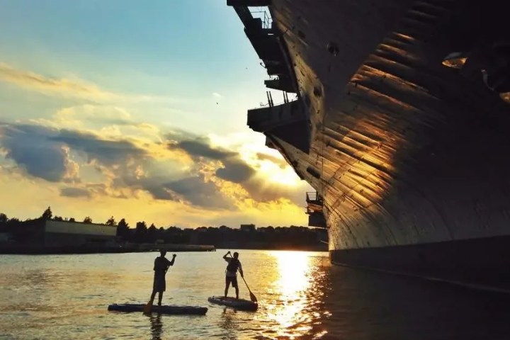Two paddleboarders silhouetted against a golden Hudson River sunset, paddling beneath the massive hull of the Intrepid at Pier 84.