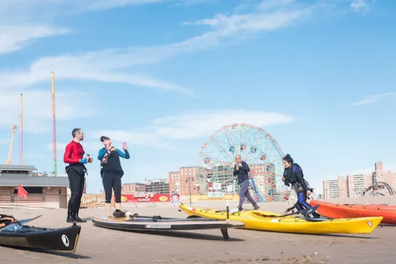 a group of people standing on the beach at Coney Island with kayaks and stand up paddle boards resting on the sand