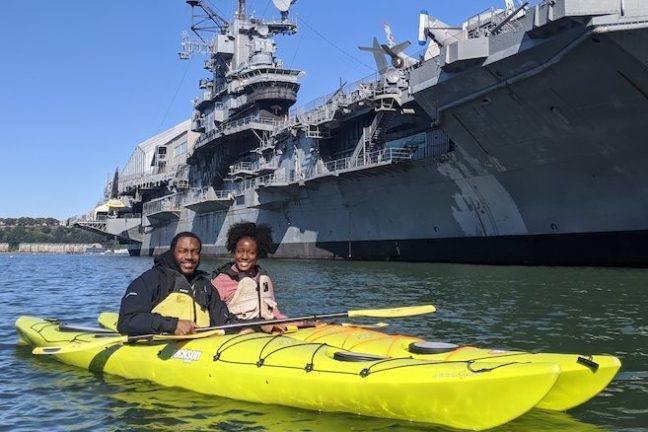 a couple in yellow kayaks in front of the Intrepid Aircraft Carrier in Manhattan