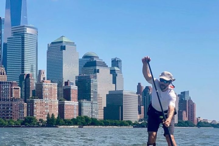 a man riding a stand up paddle board on the Hudson River with the Freedom Tower and the Financial District of Manhattan in the background