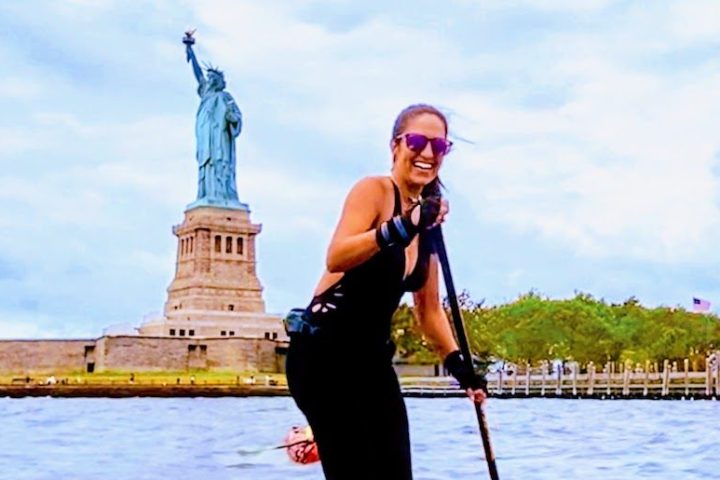 a woman wearing black on a stand up paddle board in front of the Statue of Liberty