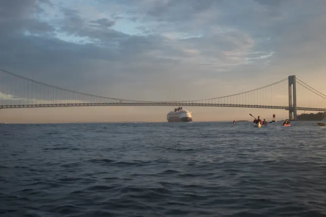 a group of kayakers and a cruise ship in front of the Verrazano Bridge in the NY Harbor during sunset