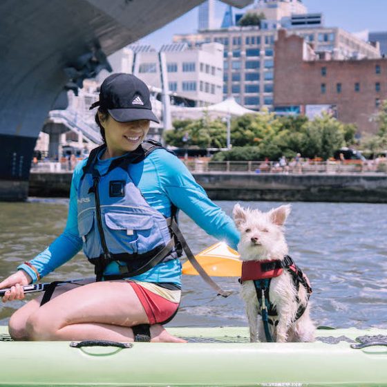 a woman on a stand up paddle board with her dog in front of the Intrepid Aircraft Carrier in Midtown Manhattan