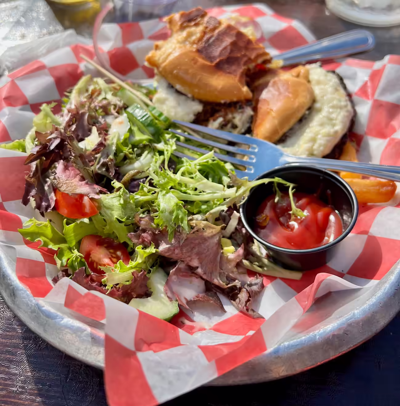 Burger and salad from the beer garden on a checkered paper-lined plate with fork and ketchup on the side.