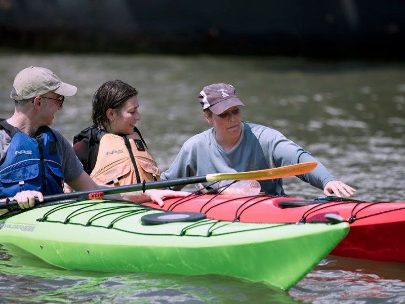 Personalized coaching in action: A senior Manhattan Kayak instructor provides immediate, hands-on feedback to a couple during a private sea kayaking lesson. They practice boat-to-body connectivity and stable strokes in colorful green and red sea kayaks in the calm Pier 84 cove.