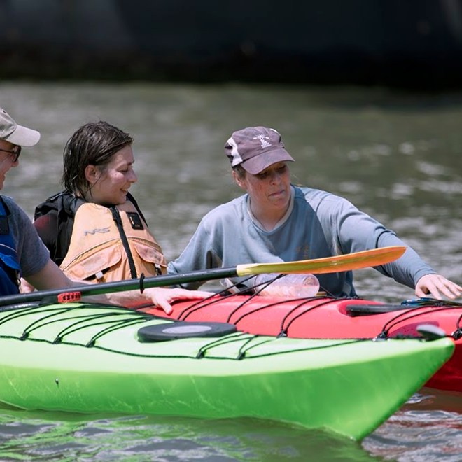Personalized coaching in action: A senior Manhattan Kayak instructor provides immediate, hands-on feedback to a couple during a private sea kayaking lesson. They practice boat-to-body connectivity and stable strokes in colorful green and red sea kayaks in the calm Pier 84 cove.