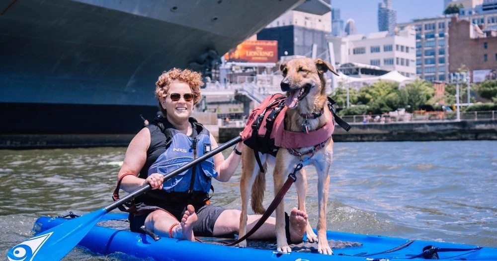 a woman and her dog on a stand-up paddleboard by the Intrepid Aircraft Carrier