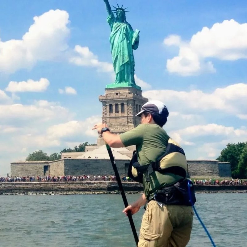An advanced paddler (Single Black Diamond) riding a paddleboard in front of the Statue of Liberty on a sunny day with crowds of people standing on Liberty Island in the distance.