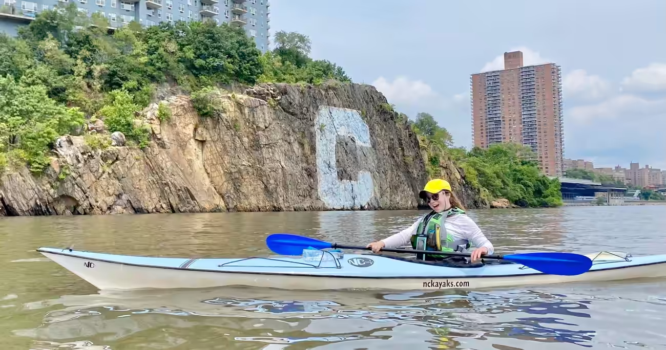 A woman riding a light blue sea kayak poses in front of the Columbia University 'C'.