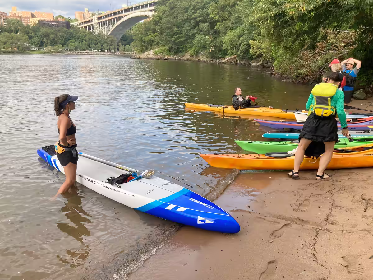 A paddleboarder standing in the water along a beach as kayaker land their kayaks.