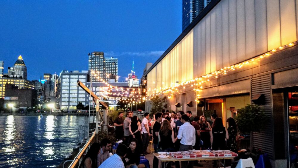 A wide shot of an outdoor patio at night with string lights, several people socializing around tables, and the Midtown skyline in the backdrop.