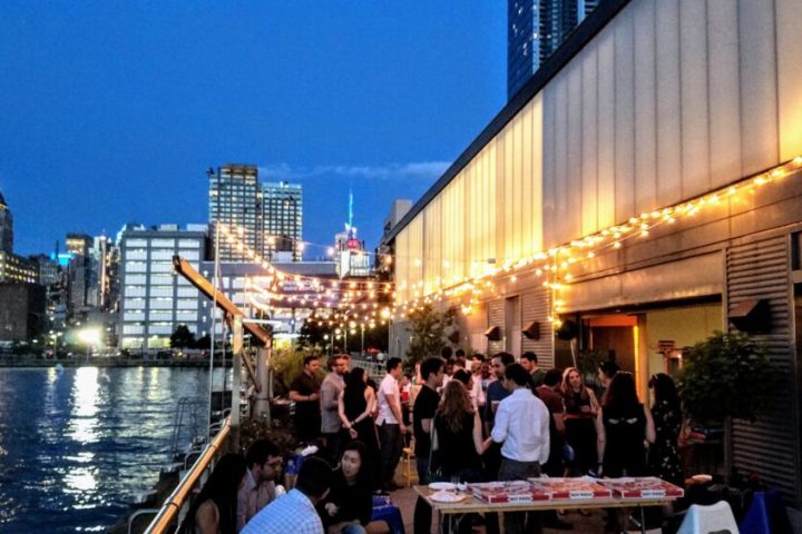 A wide shot of an outdoor patio at night with string lights, several people socializing around tables, and the Midtown skyline in the backdrop.