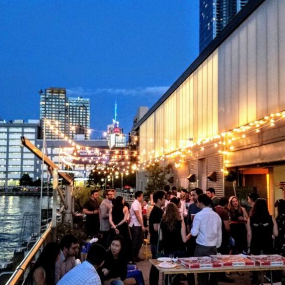A wide shot of an outdoor patio at night with string lights, several people socializing around tables, and the Midtown skyline in the backdrop.