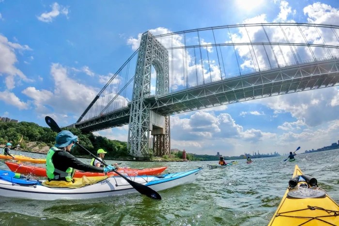 Kayakers paddling toward George Washington Bridge with the green Palisades in the background.