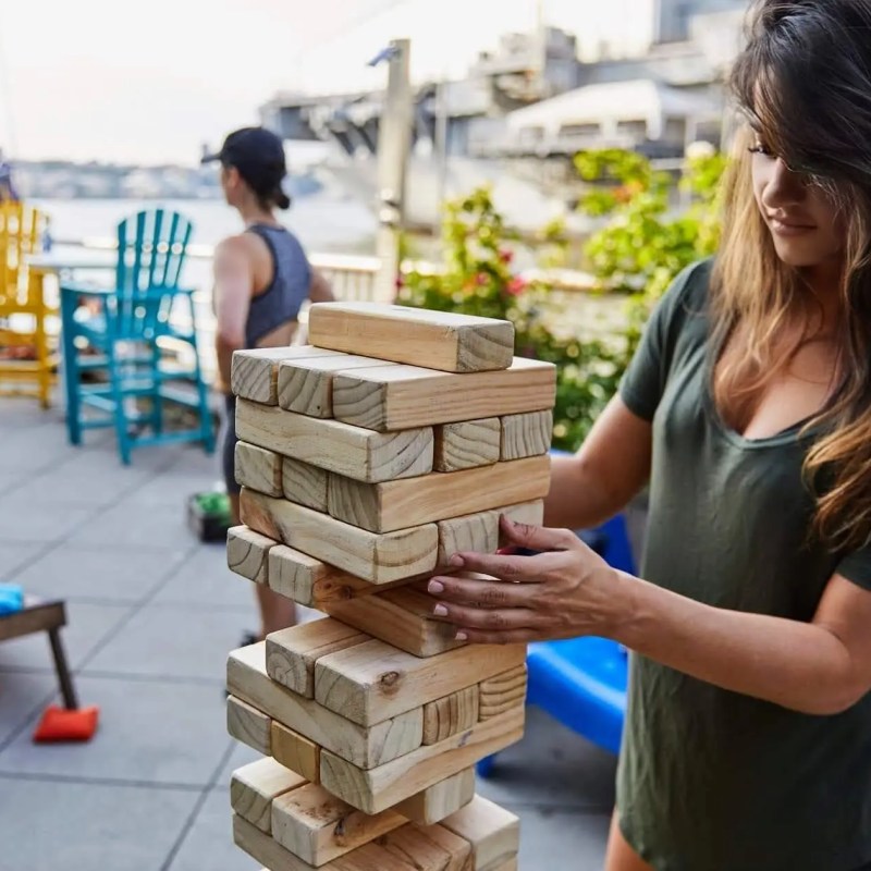 Young professionals playing giant Jenga and lawn games on an outdoor waterfront patio during a corporate summer outing at a Hudson River boathouse.