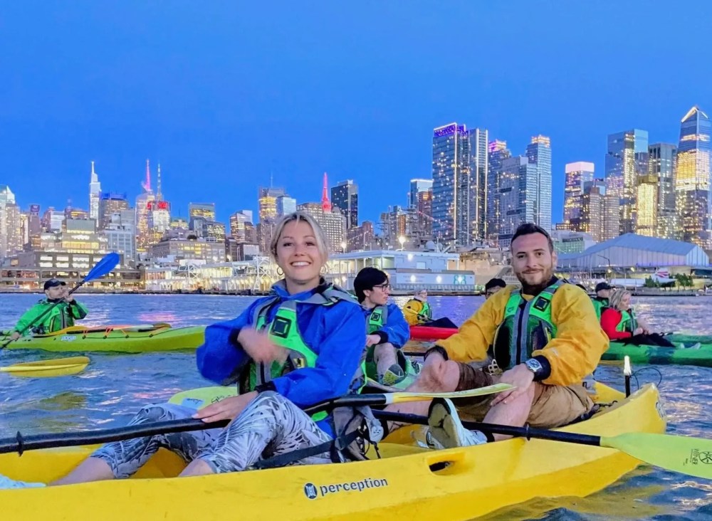 A smiling couple riding a tandem kayak during Manhattan Kayak Co's night kayaking tour on the Hudson River in NYC.