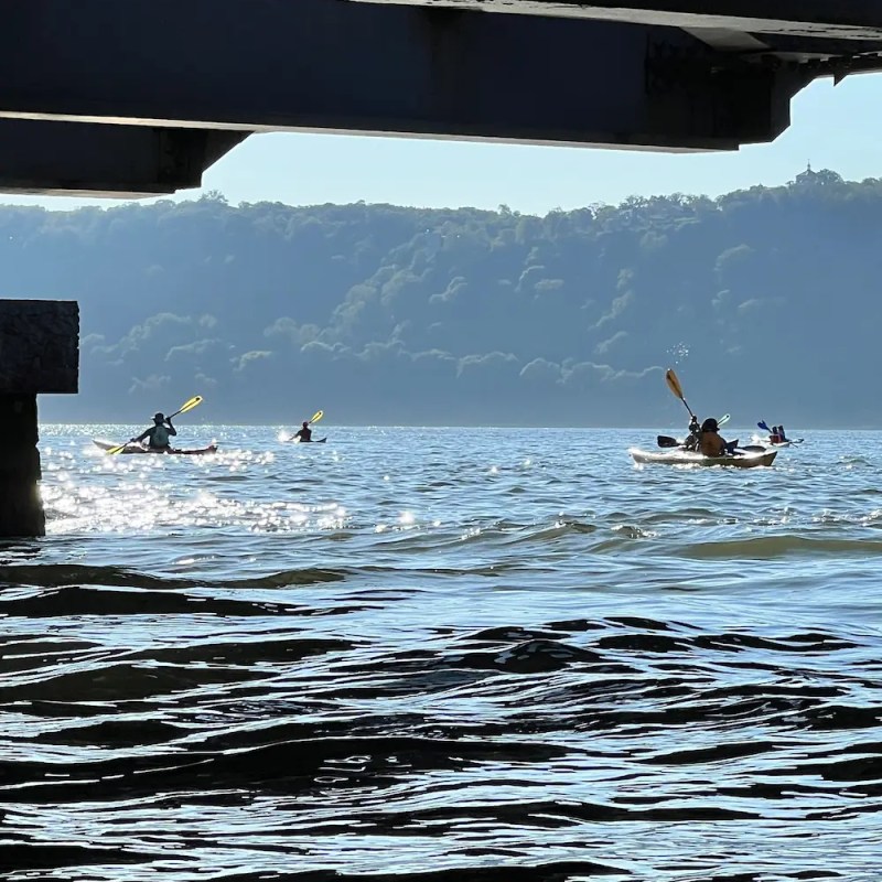 Four kayakers paddling on a calm lake under a bridge with tree-covered hills in the background.