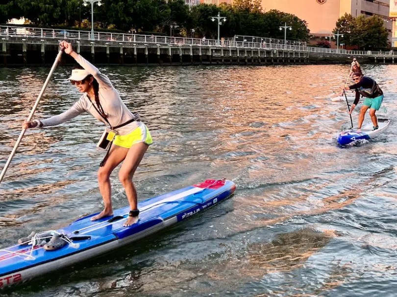 Two people paddleboarding on a river near a dock during sunset.