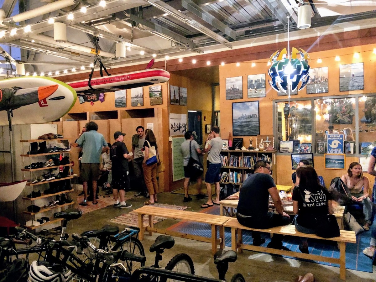 Indoor community space with people, bikes, surfboards, and wall art under string lights.