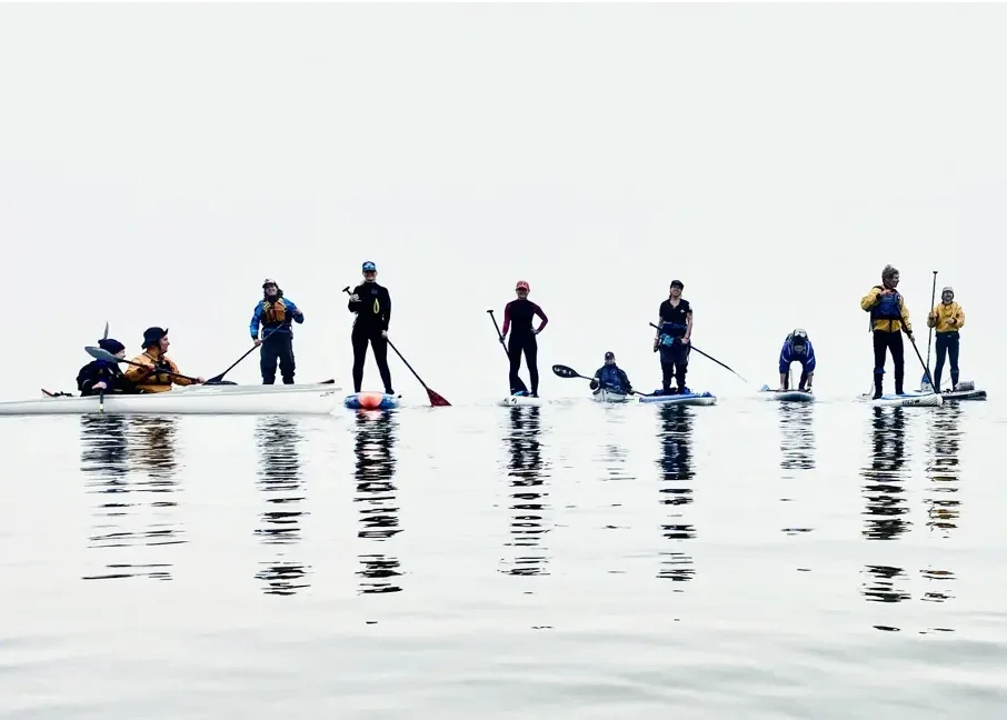 Nine Adventure Club members riding kayaks and paddlebards on calm water among solid white winter fog.