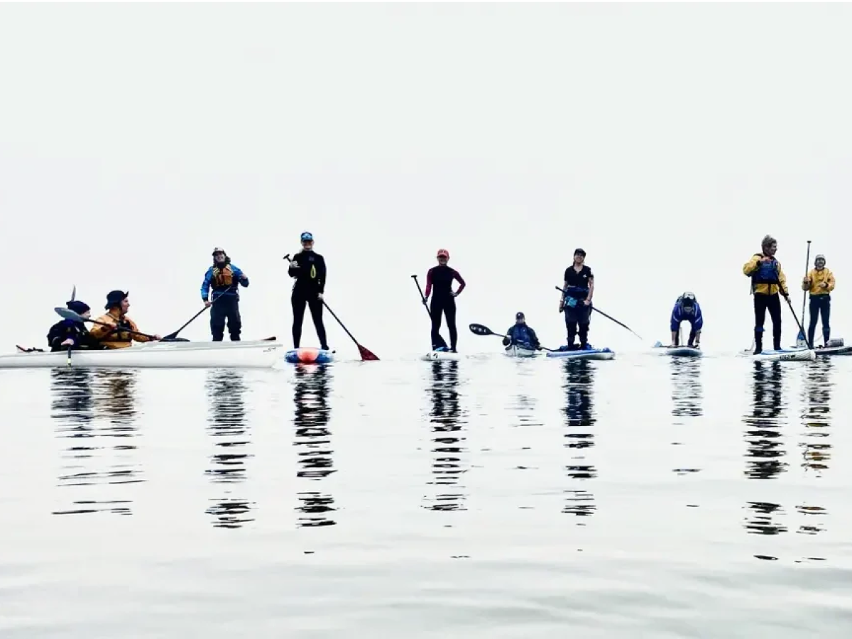 Nine people kayaking and stand-up paddling on calm water, with a clear sky background.