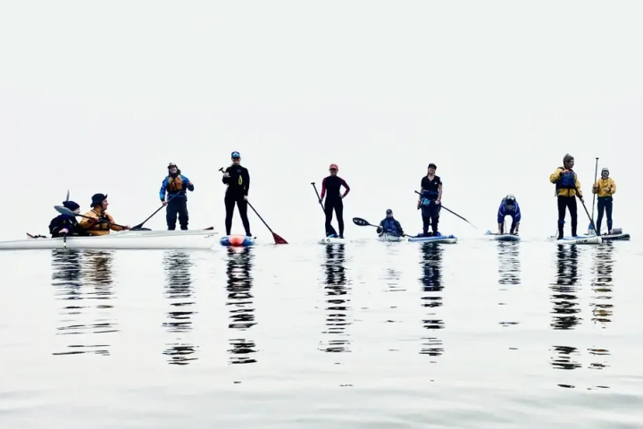 Nine Adventure Club members riding kayaks and paddlebards on calm water among solid white winter fog.
