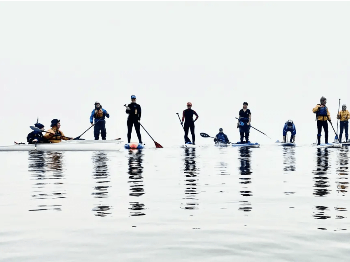 Nine people kayaking and stand-up paddling on calm water, with a clear sky background.