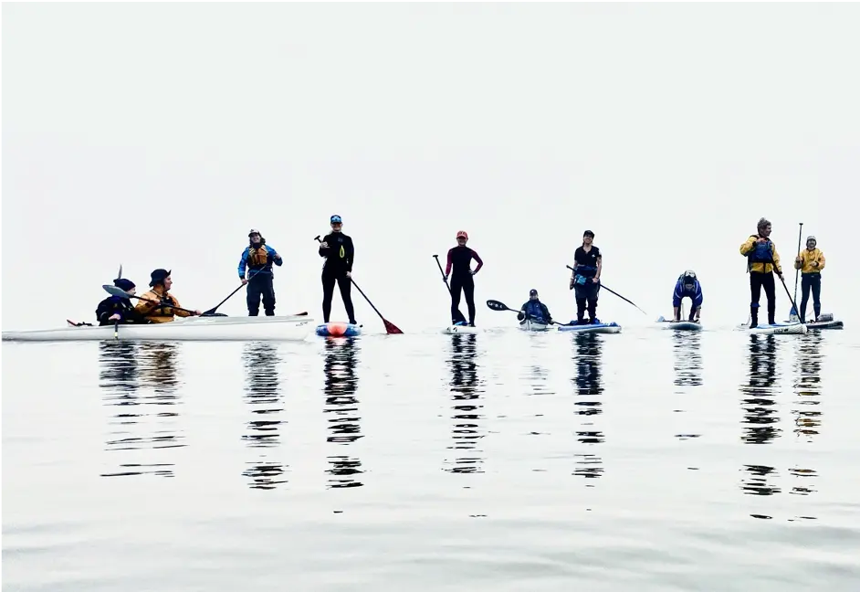 Nine people kayaking and stand-up paddling on calm water, with a clear sky background.