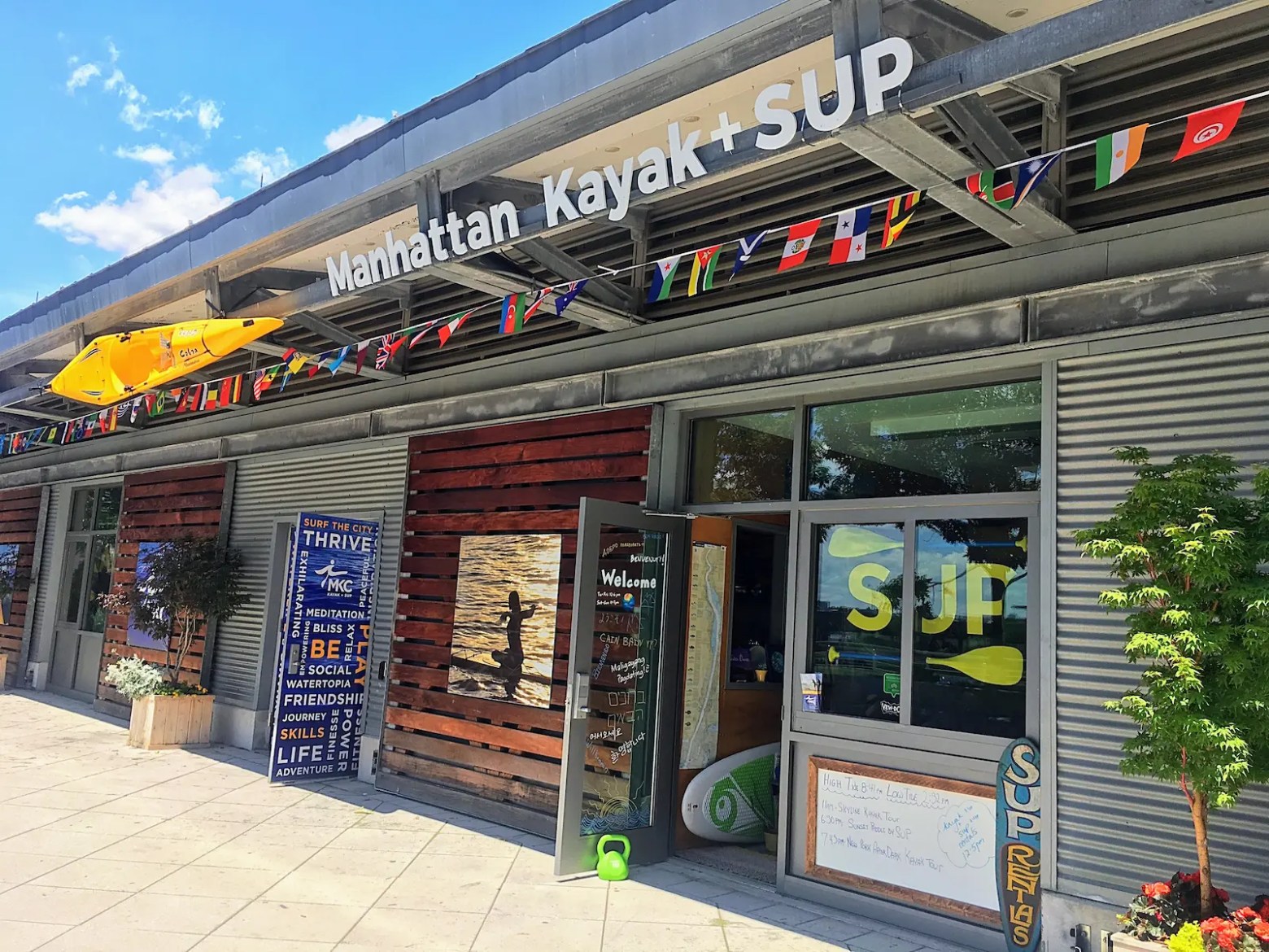 Storefront of Manhattan Kayak + SUP with flags and a yellow kayak displayed above.