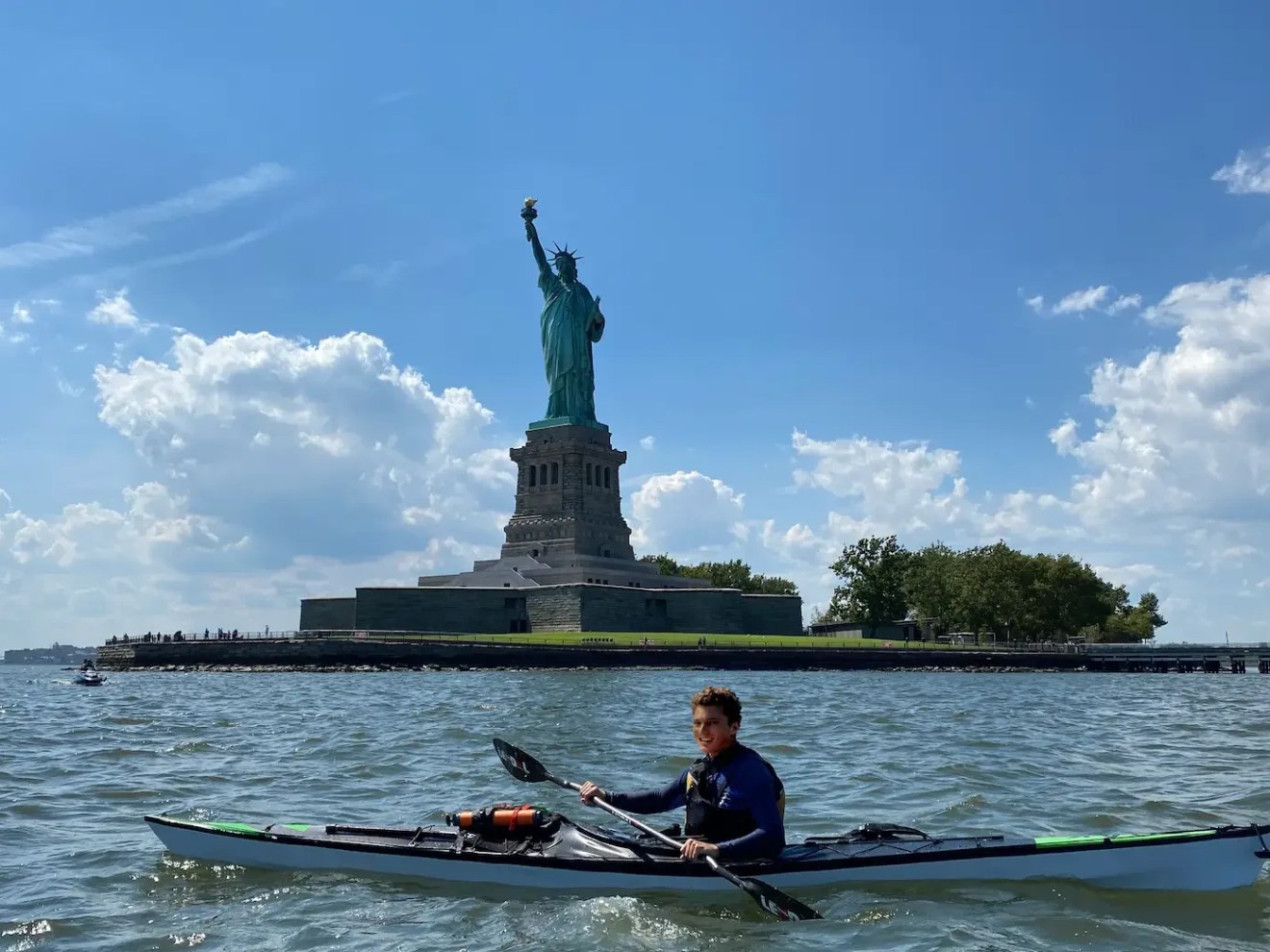 Person kayaking on water near the Statue of Liberty on a sunny day.