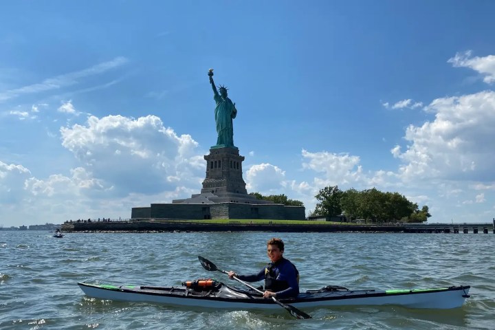 Person kayaking on water near the Statue of Liberty on a sunny day.