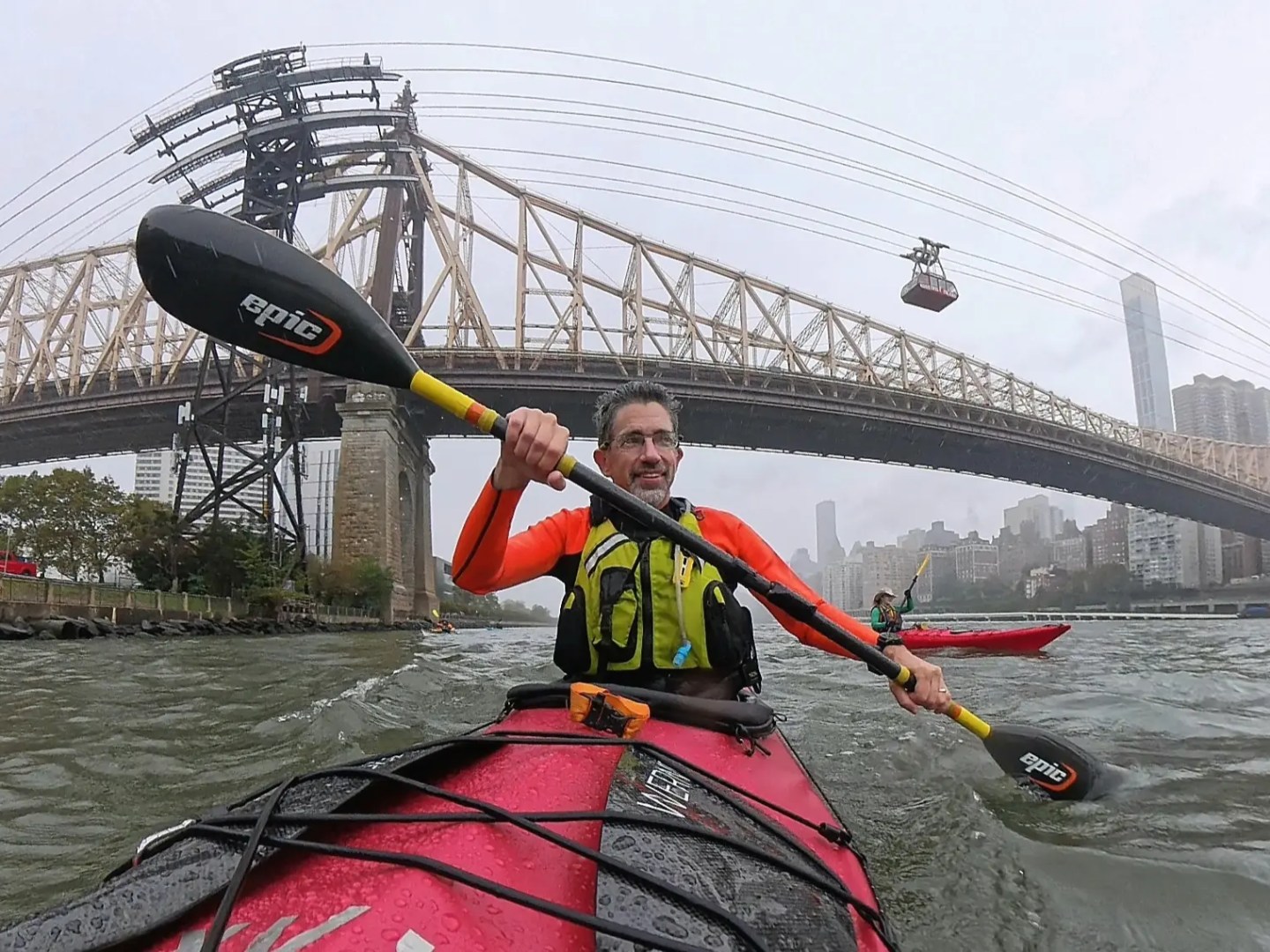 Two kayakers paddle on a river under a large suspension bridge with a cable car in the background.